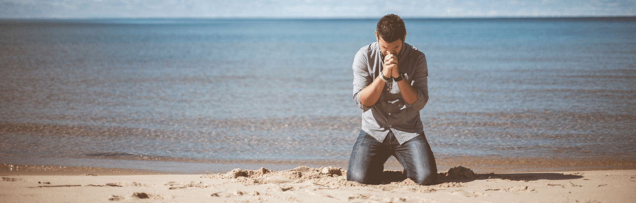 Ein Mann kniet auf dem Strand und betet, hinter ihm das Meer.